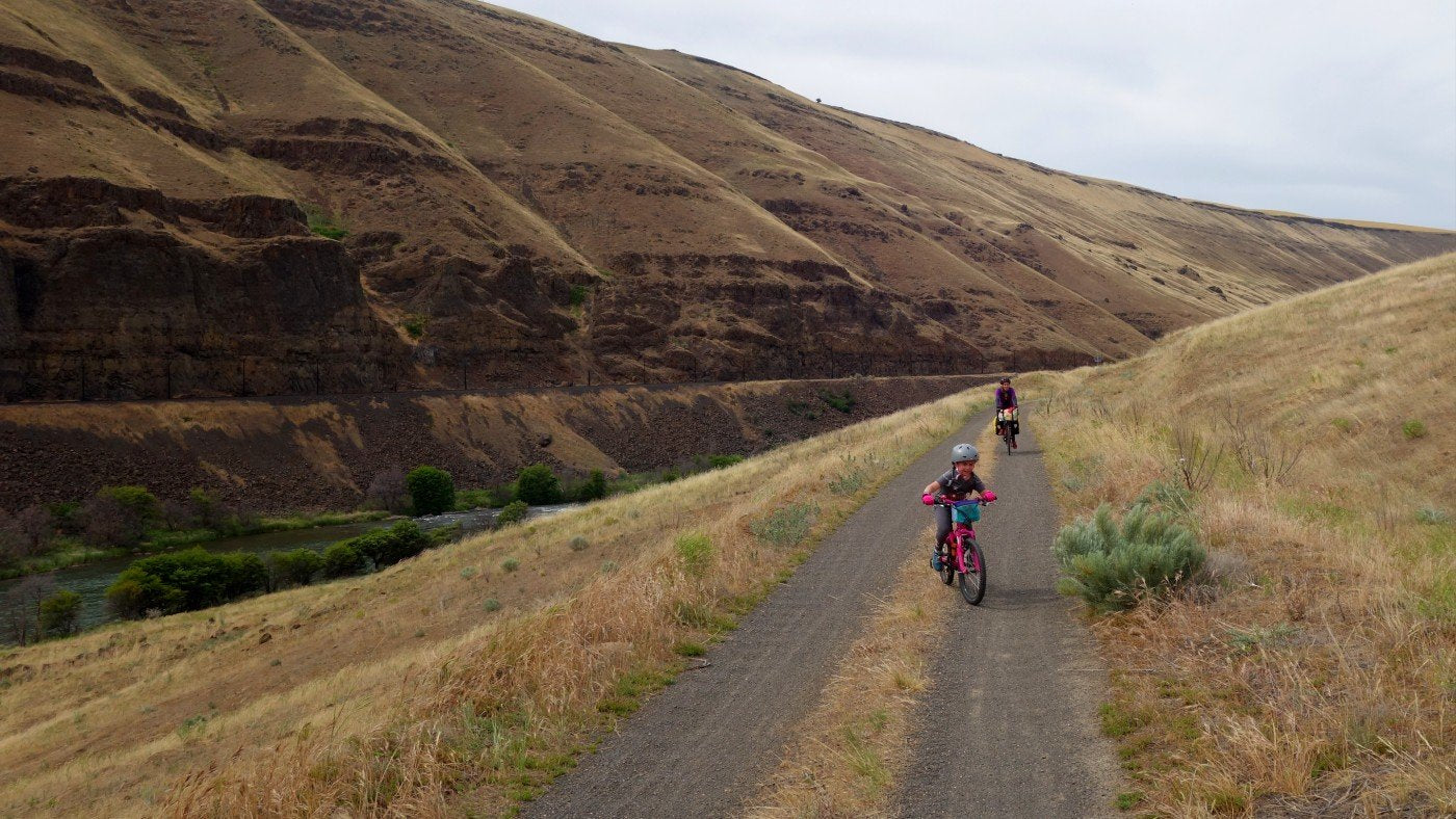 A small child on a bicycle rides a long gravel road in front of their mom on a bikepacking trip