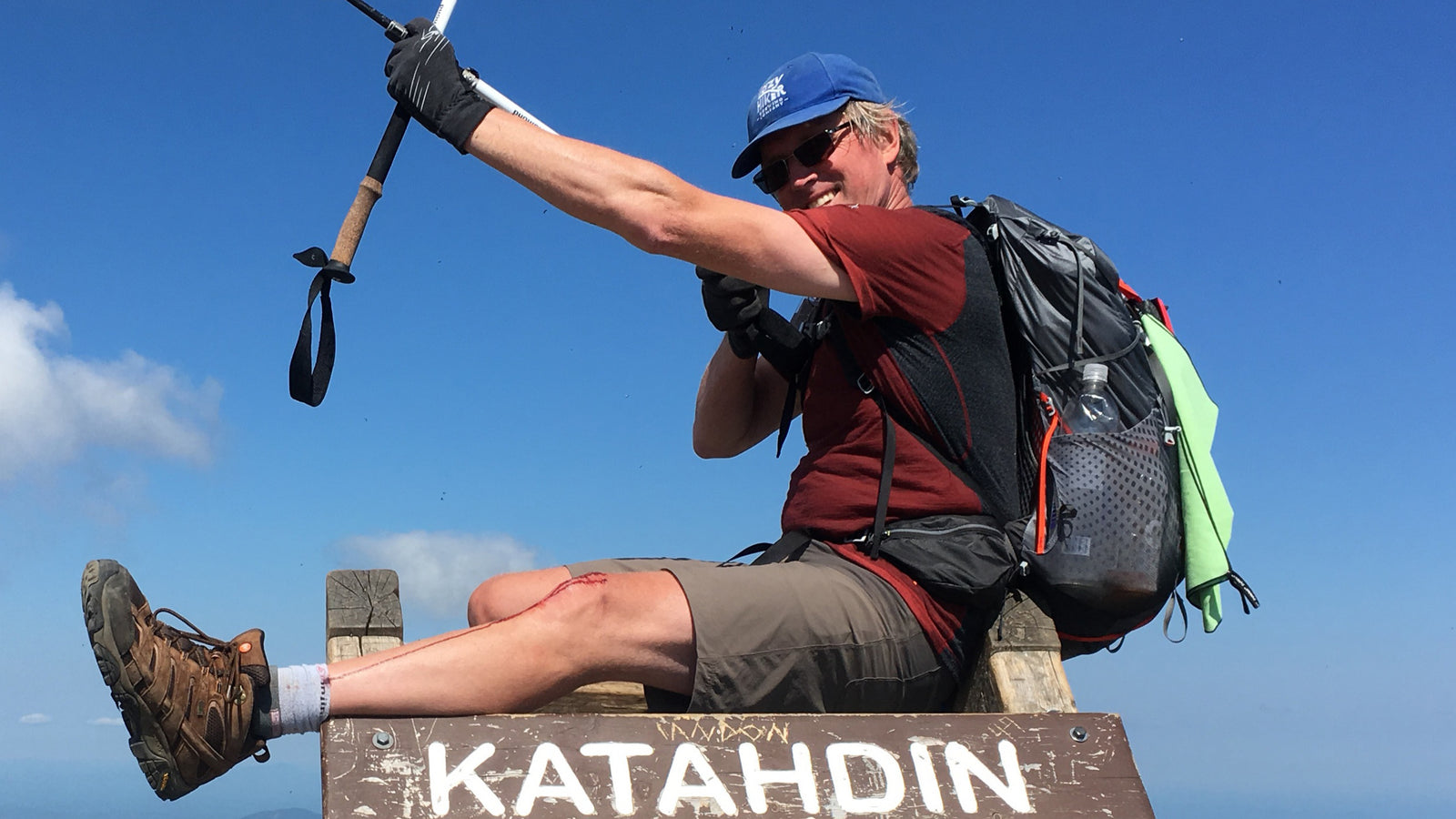 Adrian sitting on top of the Katahdin sign, smiling, with a bloody knee, holding trekking poles